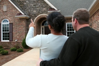 a couple stands outside of a brick home for sale, woman is pointing at the exterior