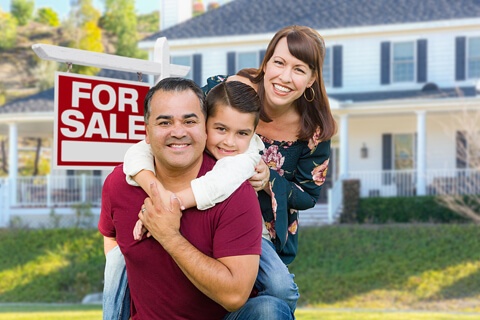 a happy family of three smiling, embracing by the FOR SALE sign in front of the new home they just purchased