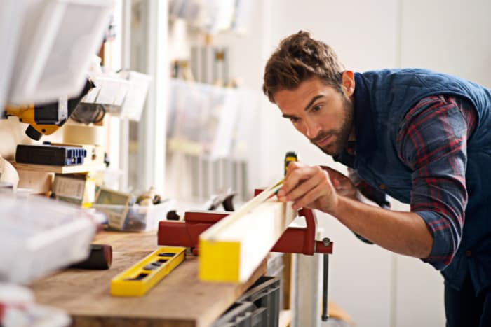 A young carpenter in flannel and a vest measuring a piece of wood.