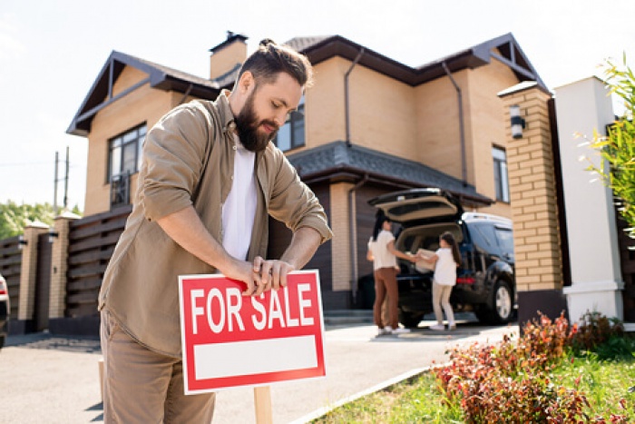 a father putting up a for sale sign in front yard, wife and child in the background packing car with moving boxes